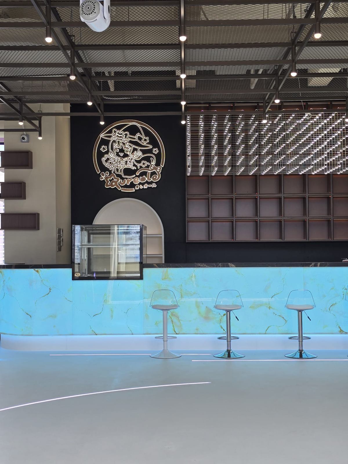 The counter lit pale blue, with three acrylic stools, a black feature wall and a steel ceiling grid.