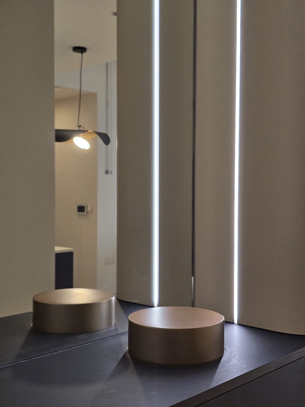 Two bronze-toned cylindrical trays on a dark console, between pale columns with lit vertical reveals.